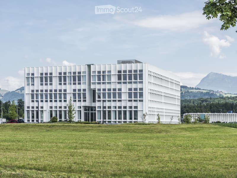 Modern, white, multi-story building with a grid of windows, surrounded by a grassy field and trees, with mountains in the background.