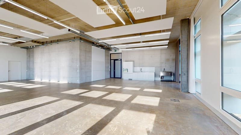 Empty room with exposed wooden ceiling beams, concrete walls, and large windows allowing natural light. Features a small kitchenette area with cabinets and a sink.