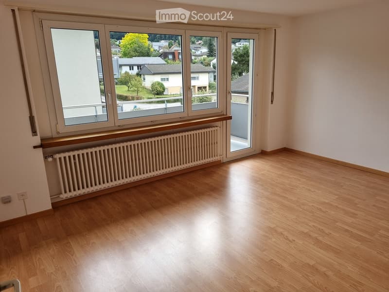 Empty room with wooden flooring, large windows, radiator, and a sliding glass door leading to a balcony.