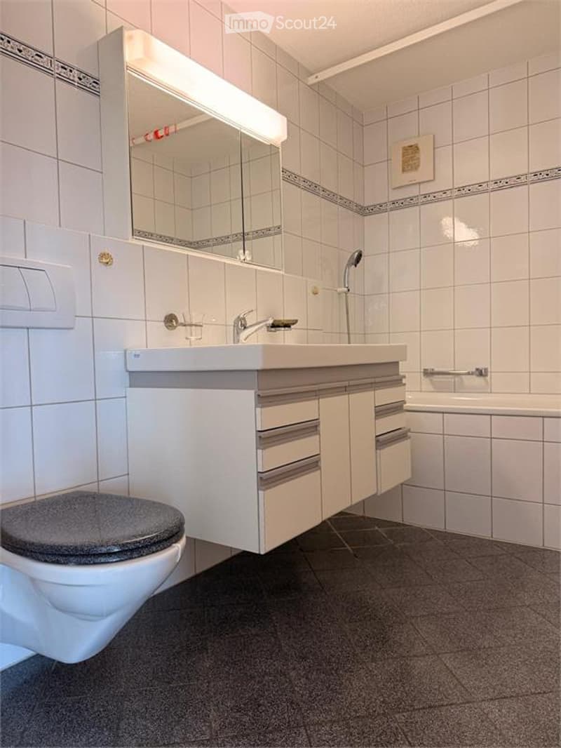 Modern bathroom with white tiled walls, a white sink with a mirror, a toilet, and a bathtub. The floor is dark gray tiles, and the space is well-lit with built-in lighting above the mirror.