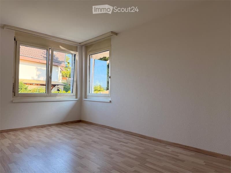 Empty room with light wooden flooring and white walls, featuring two large windows with white frames offering a view of the outside.