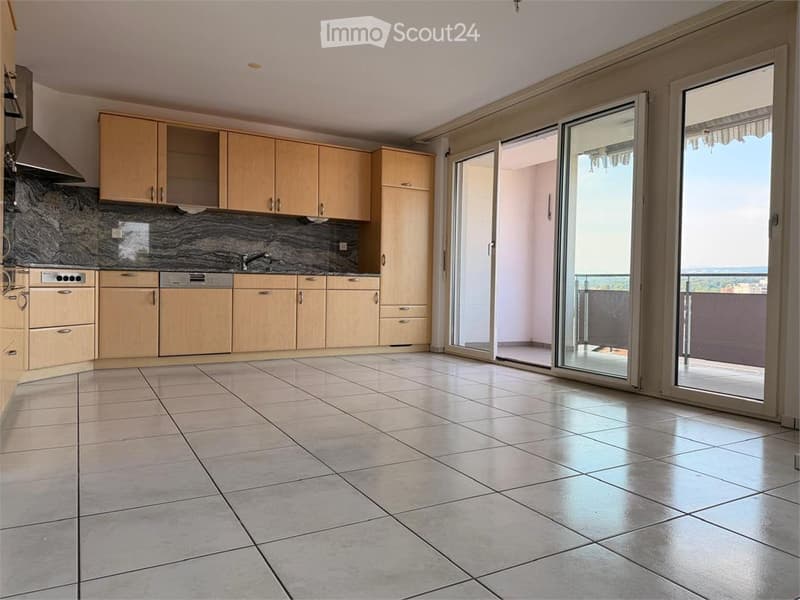 Modern kitchen with light wooden cabinets, tiled floor, and large sliding glass doors leading to a balcony.