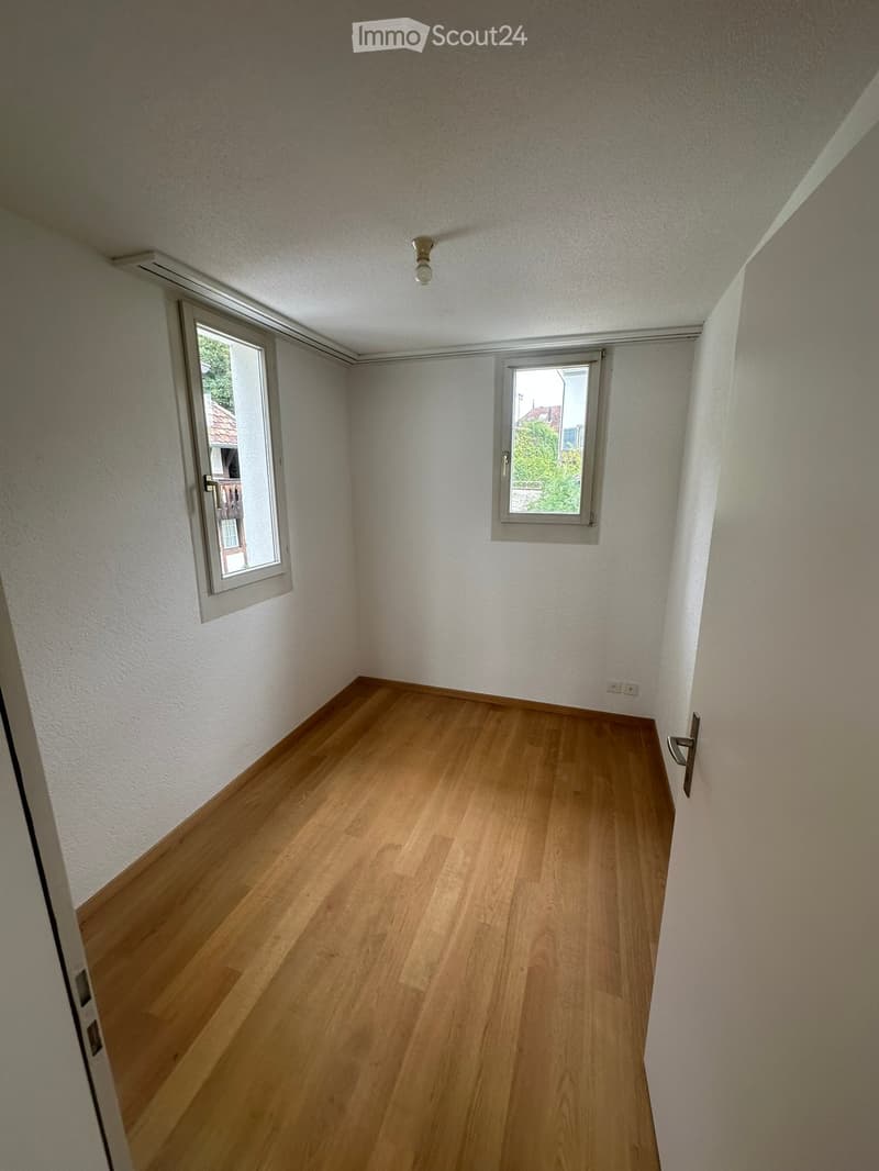 Empty room with wooden flooring, white walls, and two windows with white frames. The room has a single light fixture on the ceiling and a door on the left side.