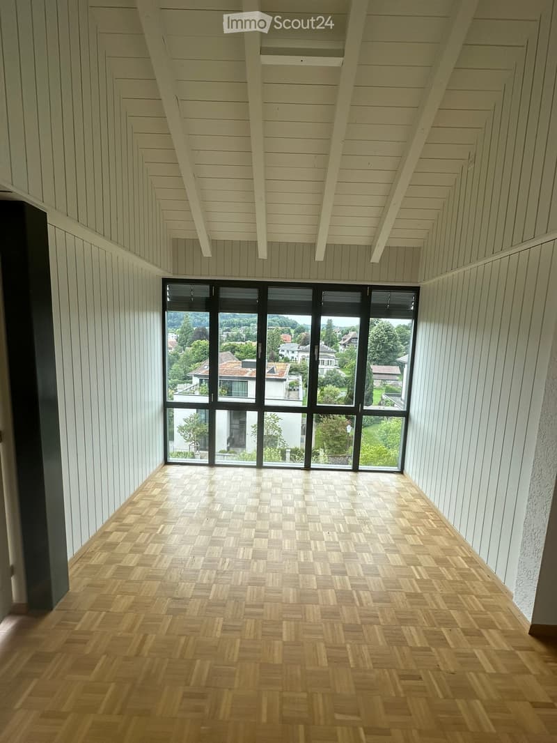 Empty room with wooden herringbone flooring, white paneled walls, and large black-framed windows overlooking a green landscape.