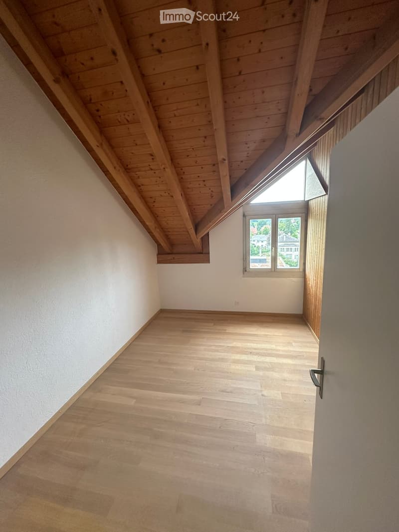 Empty room with wooden floor and slanted wooden ceiling, featuring a window with a view of the outside and a door.