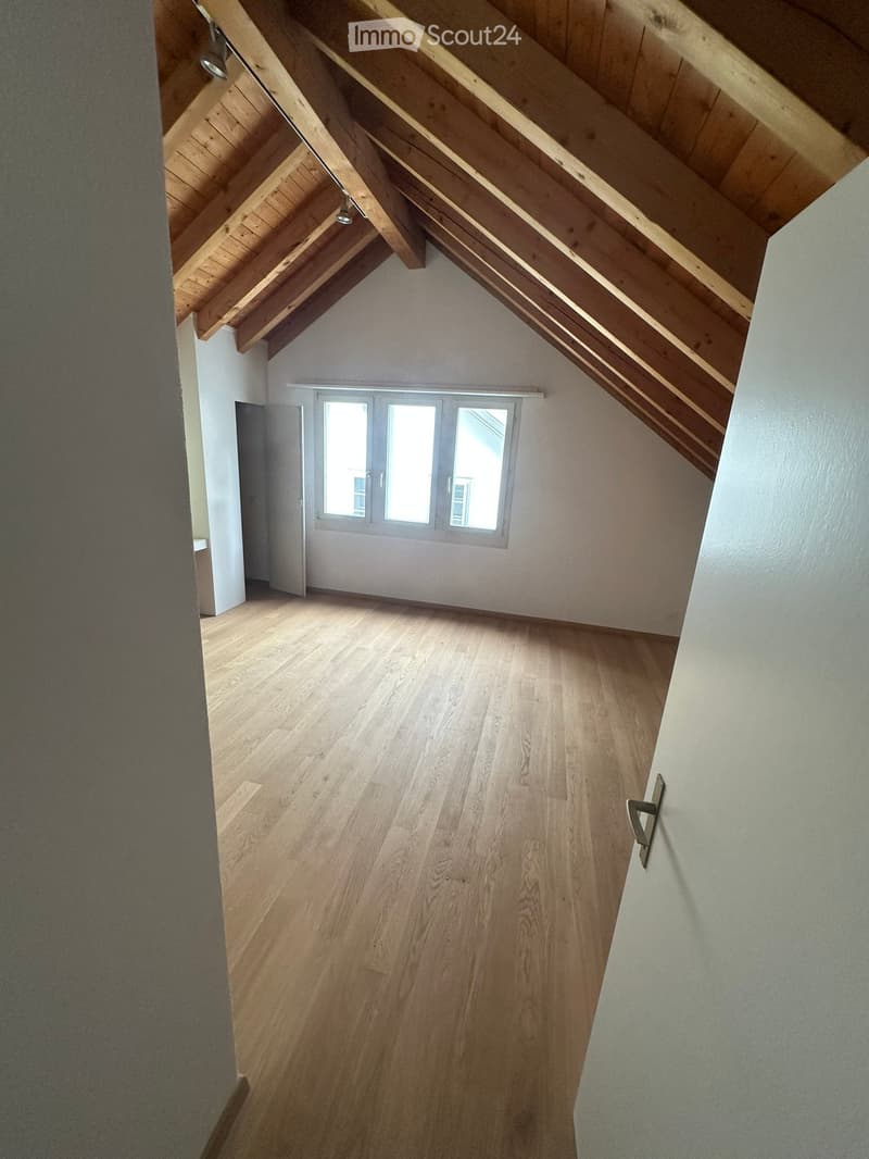 Empty attic space with wooden slanted ceilings, light wooden flooring, and large windows allowing natural light.