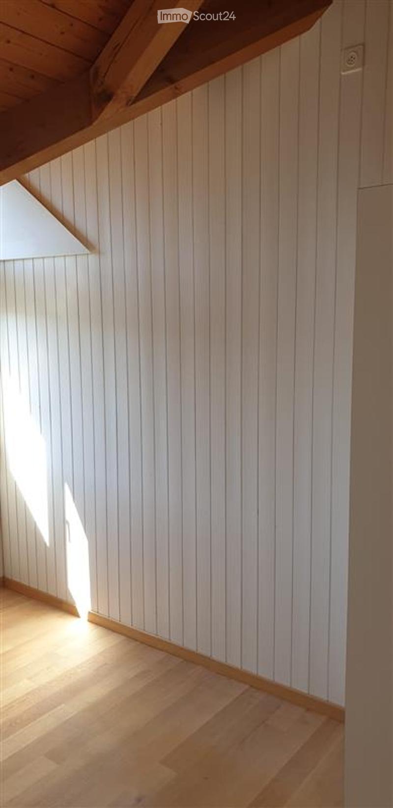 An empty room with white paneled walls and light wooden flooring, featuring a slanted wooden ceiling and a small window allowing natural light to enter.