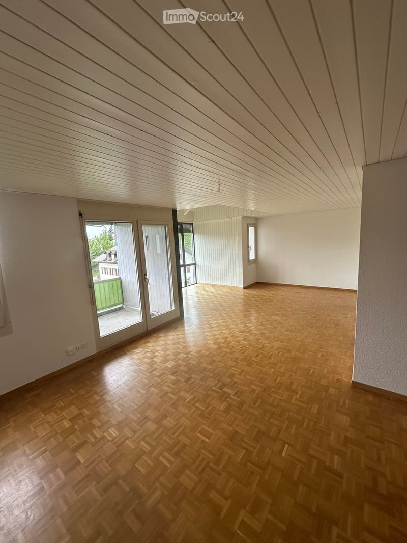 Empty room with wooden herringbone flooring, white walls, and a slanted wooden ceiling. Features large sliding glass doors leading to a balcony.
