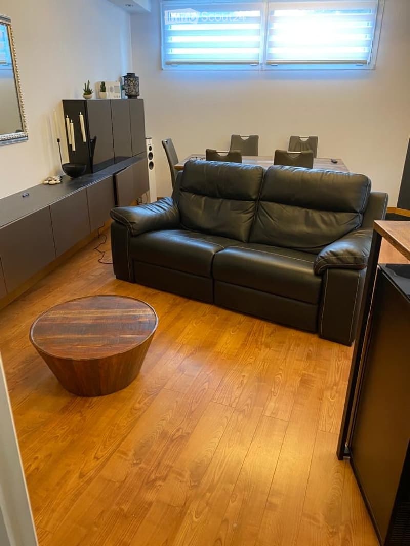 Modern living room with hardwood flooring, a black leather sofa, a round wooden coffee table, a dining area with a table and chairs, and a sleek cabinet with a mirror and decorative items.