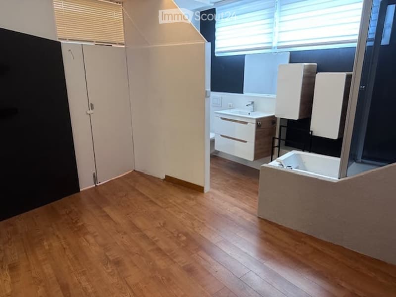 Empty bathroom with wooden flooring, white walls, and a large window with blinds. The space includes a sink, a bathtub, and a mirrored cabinet, but no furniture or personal items are present.