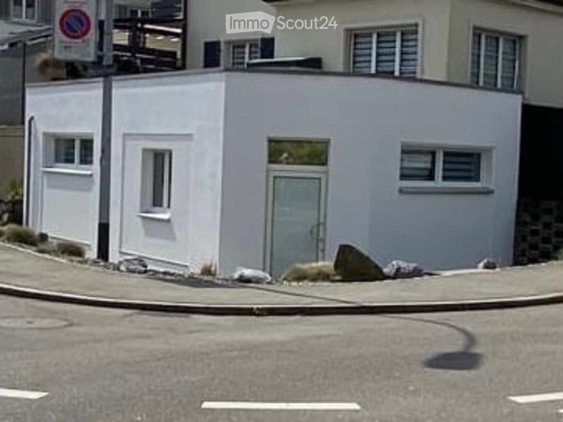 A small, modern, white building with a flat roof and large windows, situated on a corner lot with a paved street in front.