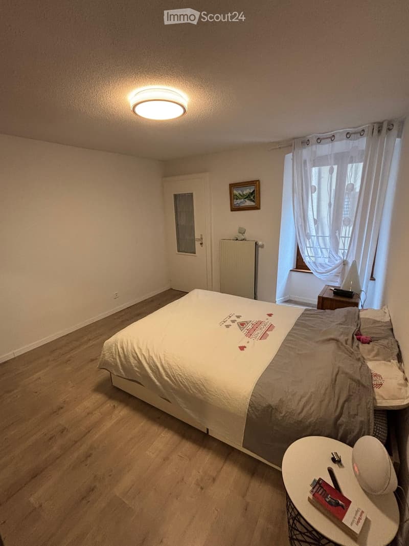 Cozy bedroom with wooden flooring, a double bed with patterned bedding, a nightstand, a small round table with items, a radiator, and a framed picture on the wall.