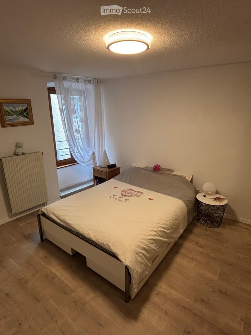 Cozy bedroom with a double bed, nightstand, small table, radiator, and a window with curtains.