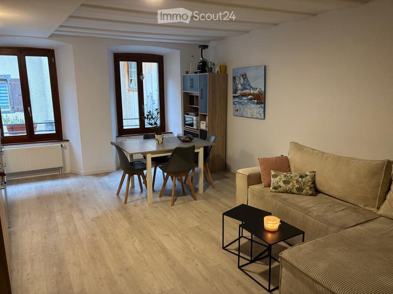 Modern living and dining area with a sofa, coffee table, dining table, chairs, and a small cabinet. The space features wooden flooring and large windows with a view.
