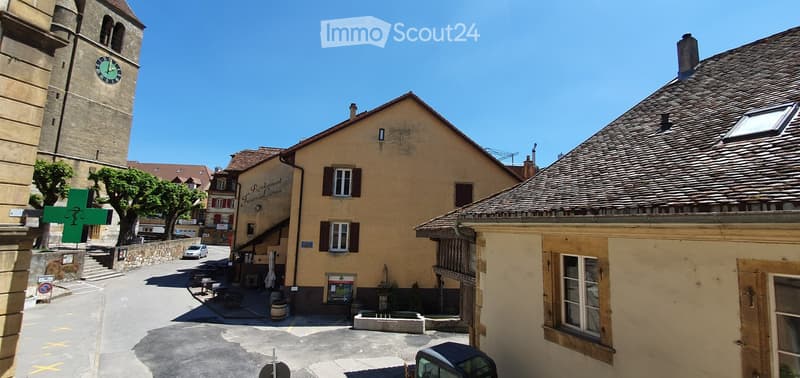 A picturesque European town square with a tall clock tower, traditional stone buildings, a restaurant with outdoor seating, and a clear blue sky.
