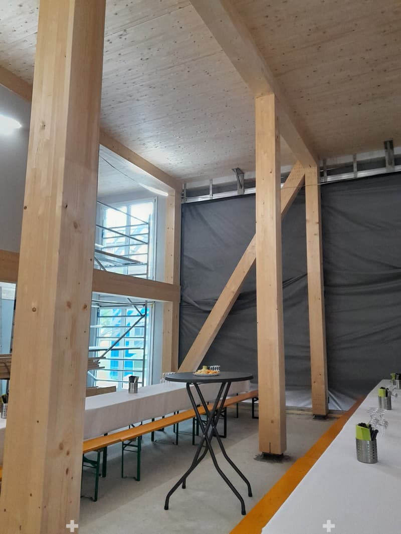 Modern, open-concept space with exposed wooden beams and pillars, featuring a long bench seating area, a small round table, and a bar area with wine glasses and utensils.