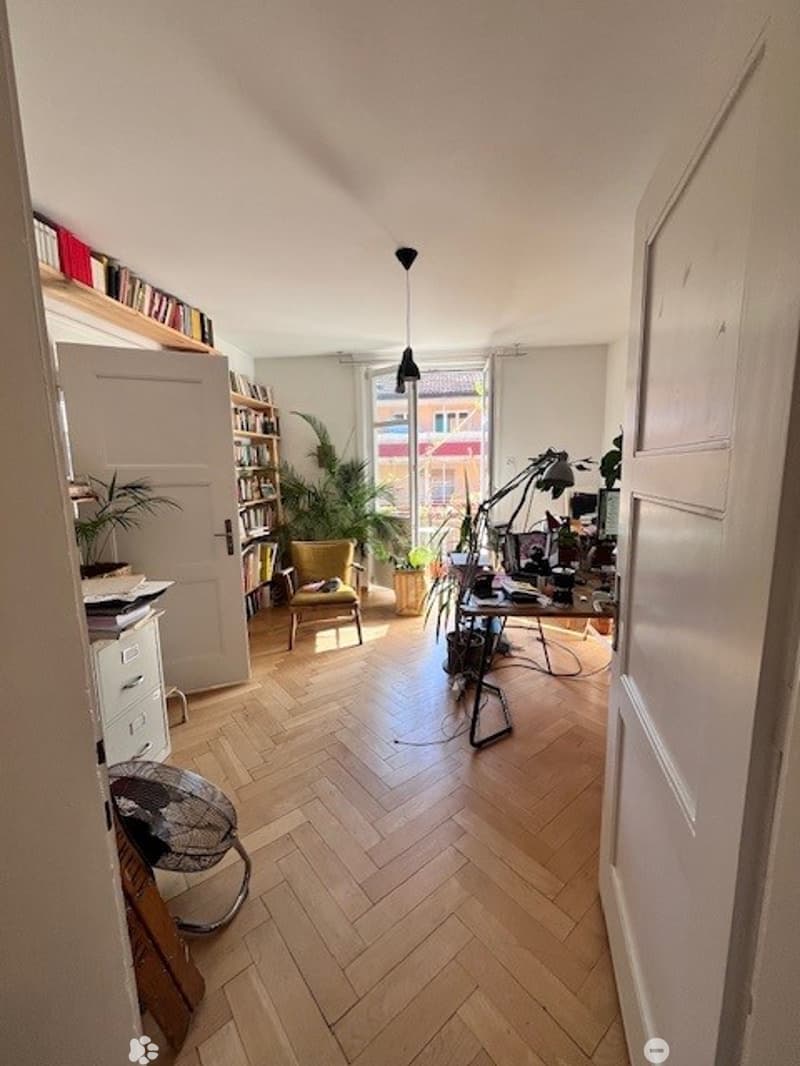 A well-lit room with herringbone wooden flooring, a large window, a desk with a computer, a bookshelf, a chair, and a small fan. The room is decorated with plants and has a cozy, organized feel.