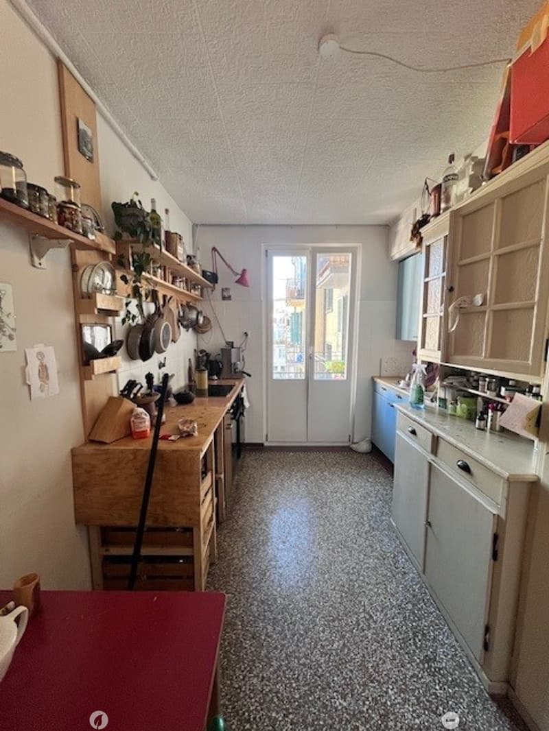 A cozy kitchen with wooden countertops, pantry shelves filled with pots and pans, and various kitchen utensils. The space includes a sink, a fridge, and a small balcony visible through the door.