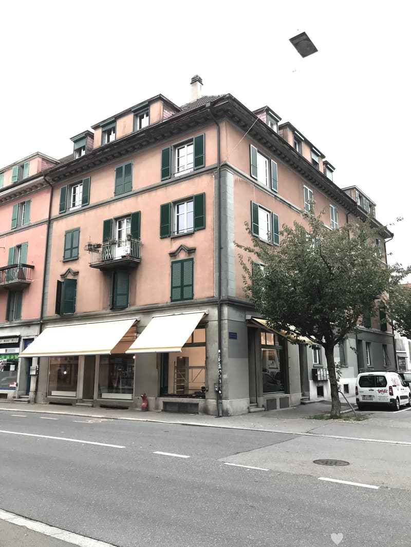 A four-story apartment building on a street corner with multiple windows and balconies on all levels. The facade is beige with large green shutters. There is an awning in front of the building, and a fire extinguisher is placed next to it. There are two t