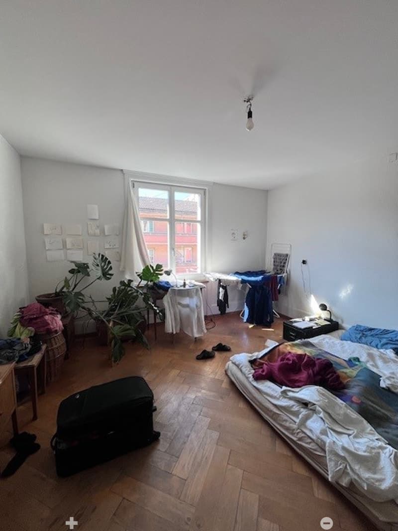 A small, lived-in bedroom with a wooden floor, a bed with colorful bedding, a small table with a plant, a suitcase, and a lamp on a nightstand.