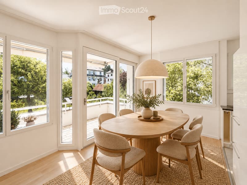 Bright dining area with a round wooden table surrounded by six upholstered chairs, a large pendant light, and a vase with greenery on the table. Large windows and sliding doors open to a balcony with greenery visible outside.