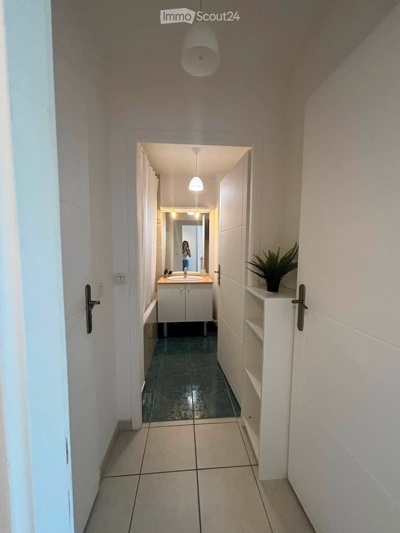 A narrow hallway with tiled floors, white walls, and a small bathroom visible at the end. The bathroom features a sink, mirror, and a small potted plant on a shelf.
