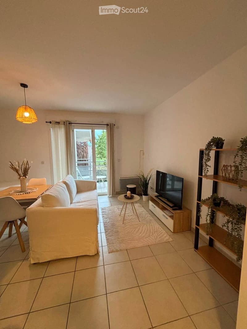Modern living room with a white sofa, coffee table, TV stand, and a small dining area. Features a balcony with outdoor furniture visible through large glass doors.