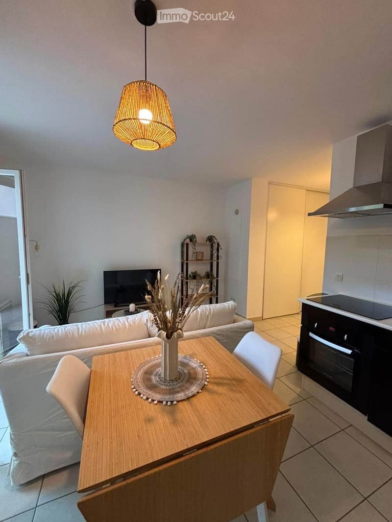 A modern, open-concept living and dining area with a white sofa, wooden dining table, chairs, a vase with dried flowers, a television, and a small kitchen area with an oven and shelf.