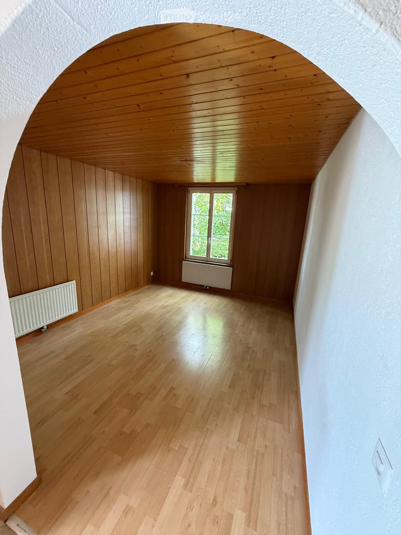 Empty room with wooden flooring, wooden walls, and a large window with a radiator below it. The ceiling is also wooden with a curved design.