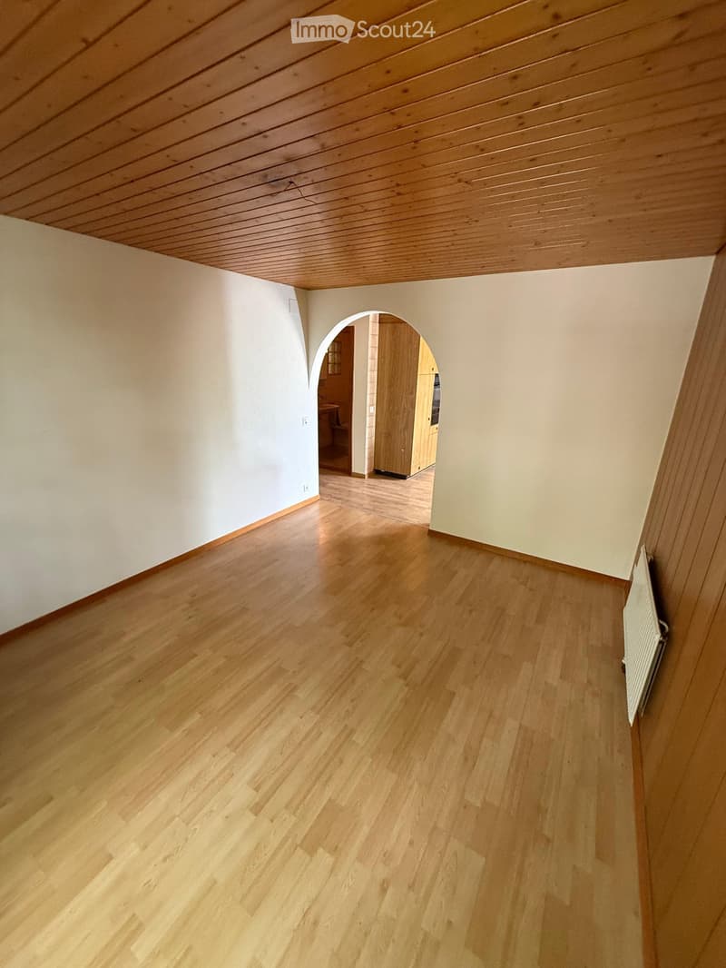 Empty room with wooden flooring, white walls, and a slanted wooden ceiling. An arched doorway leads to another room with visible furniture.