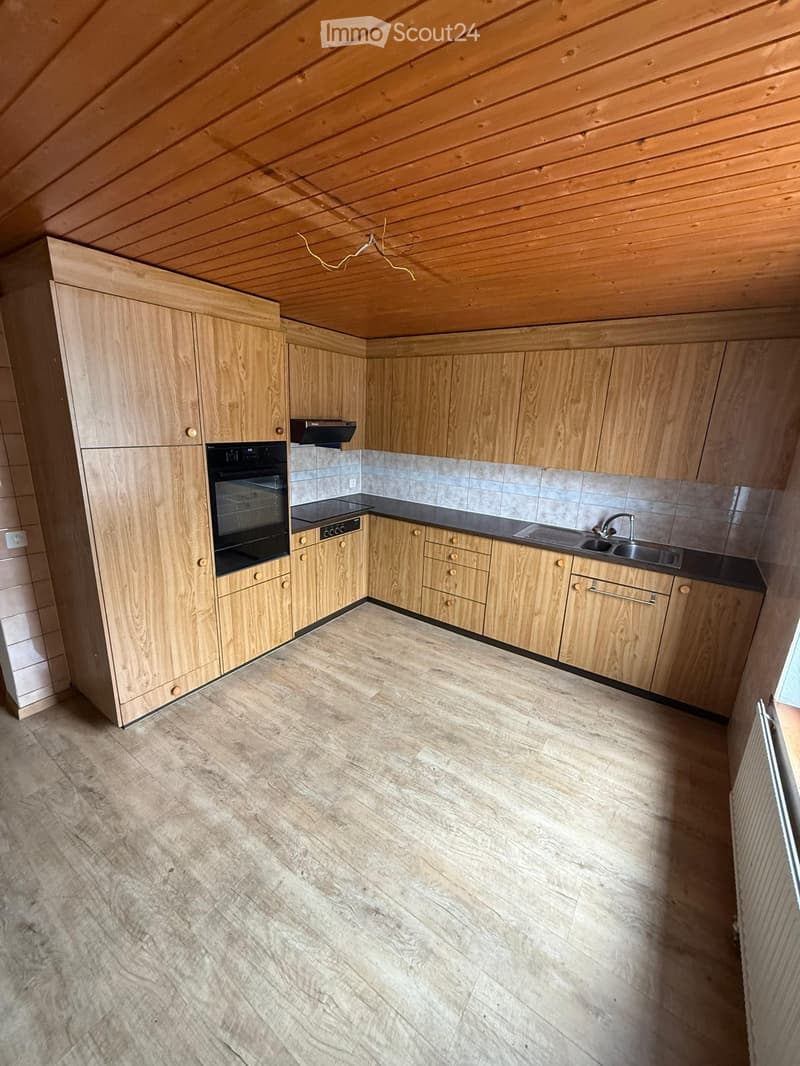 Empty kitchen with light wooden cabinets, stainless steel sink, and tiled backsplash. The floor is light-colored wood laminate.