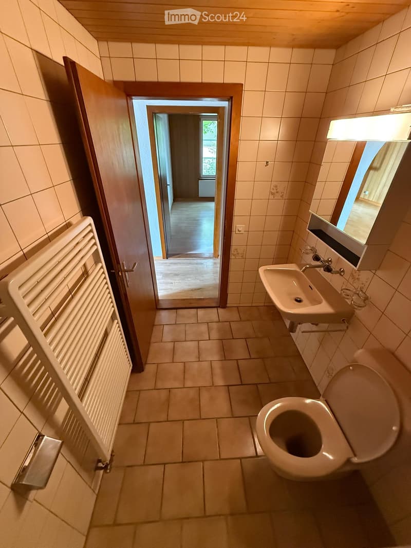 Bathroom with tiled walls and floor, featuring a toilet, sink, and radiator. A doorway leads to another room with wooden flooring.