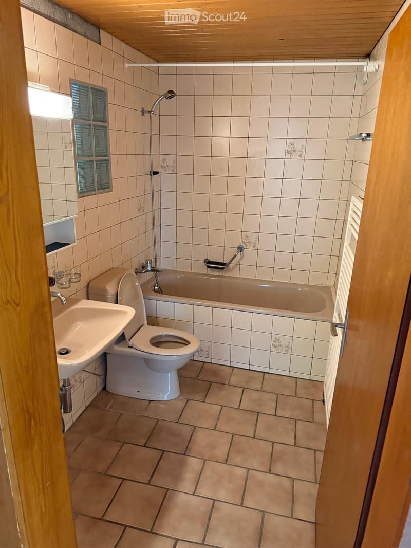 Bathroom with beige tiles, a toilet, a bathtub, a sink, and a showerhead. The room has a wooden ceiling and a small window with frosted glass.