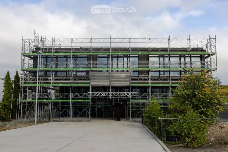 Modern multi-story building under construction with scaffolding, large glass windows, and green accents on the facade.