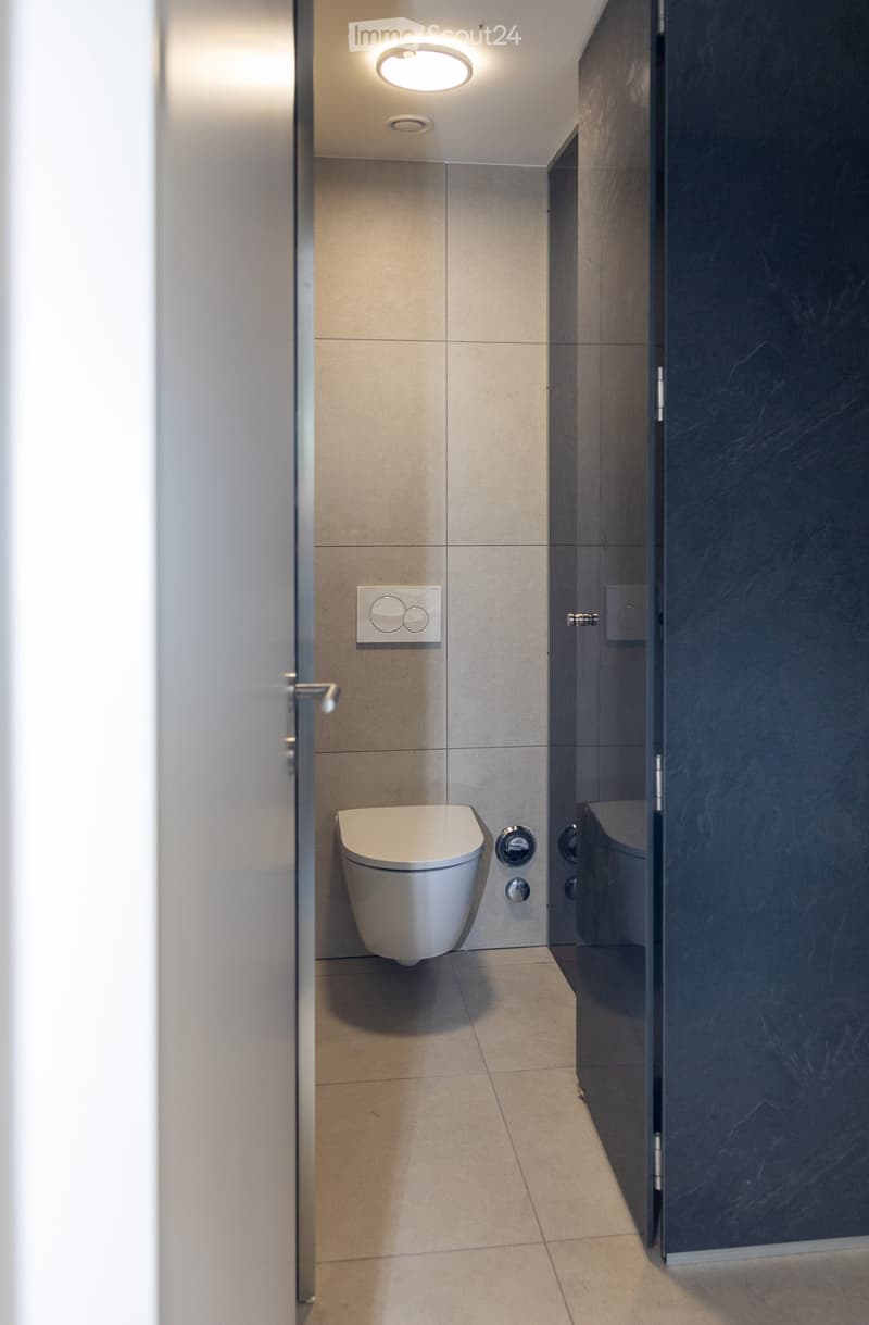 Modern bathroom with beige tiled walls and floor, a white toilet, and a dark grey shower enclosure with glass door.