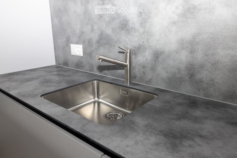 Modern kitchen sink with a sleek stainless steel faucet and a minimalist design, set against a grey countertop and textured wall.
