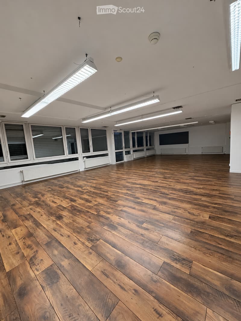 Empty room with wooden flooring, large windows, and fluorescent lighting fixtures.
