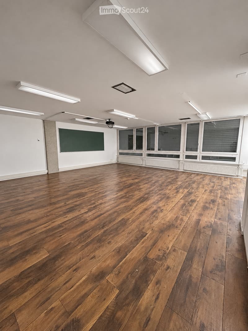 Empty room with wooden flooring, white walls, large windows, and a dark green chalkboard on one wall.