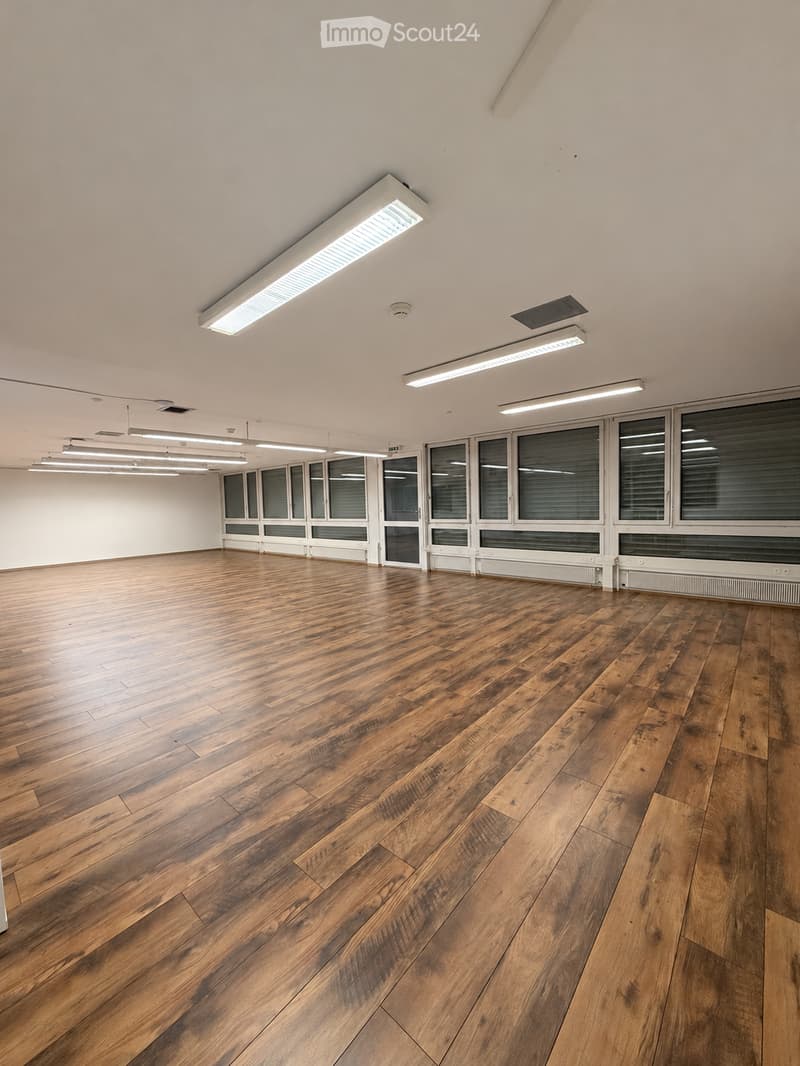 Empty room with wooden flooring, large windows, and bright overhead lighting.