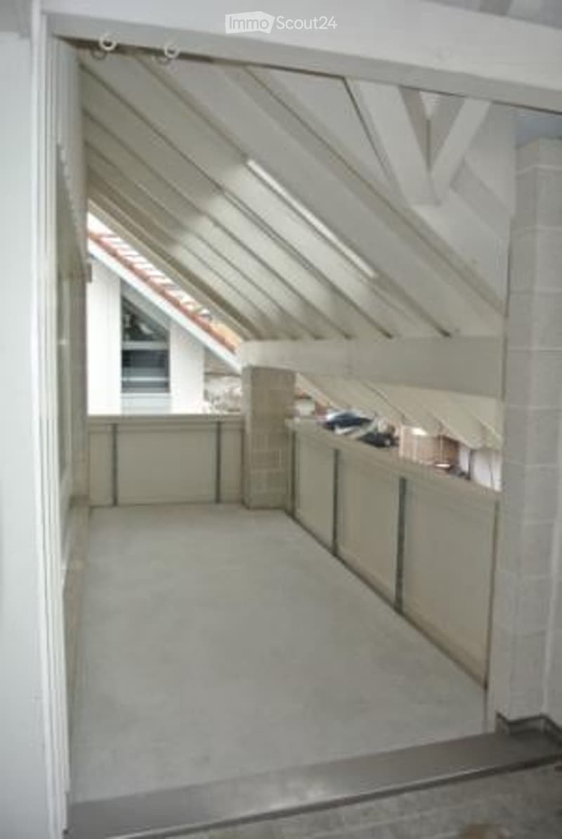 A narrow, well-lit hallway with white walls, a concrete floor, and a slanted ceiling with exposed beams. The hallway leads to a small open area with a view of another building through a window.