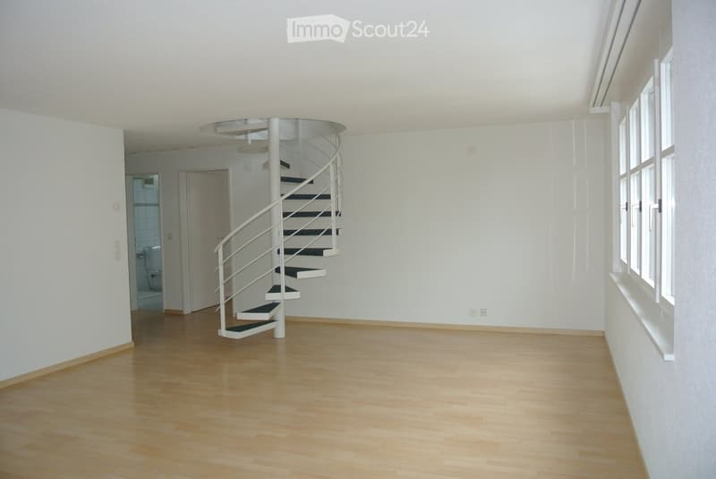 Empty room with light wooden flooring, white walls, and a modern spiral staircase leading upwards. The room has large windows on one side and a doorway leading to another area.