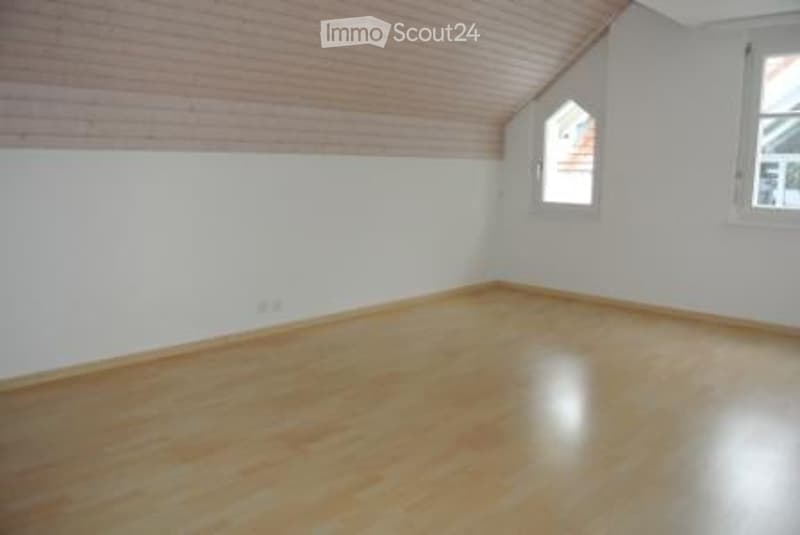 Empty room with wooden flooring, white walls, and two windows, one of which is a dormer window with a triangular shape.