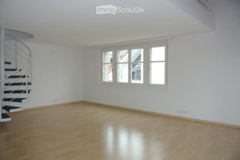 Empty room with wooden flooring, white walls, and large windows. A spiral staircase is visible on the left side.