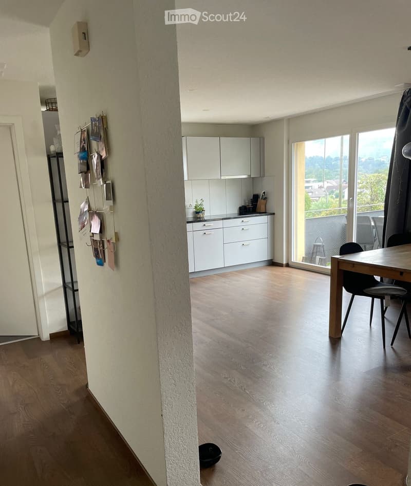 Open-plan living and dining area with light wood flooring, white kitchen cabinets, a dining table with chairs, and a view of a balcony.