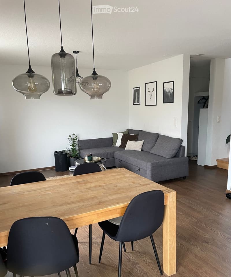 Modern living and dining area with a grey sectional sofa, wooden dining table with four chairs, pendant lights, and decorative picture frames on the wall.