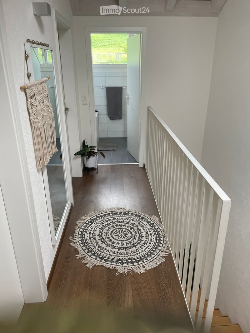 A modern hallway with wooden flooring, white walls, a white railing, and a round decorative rug in the center. A small plant and a macrame wall hanging are visible on the left side, and a bathroom is visible at the end of the hallway.