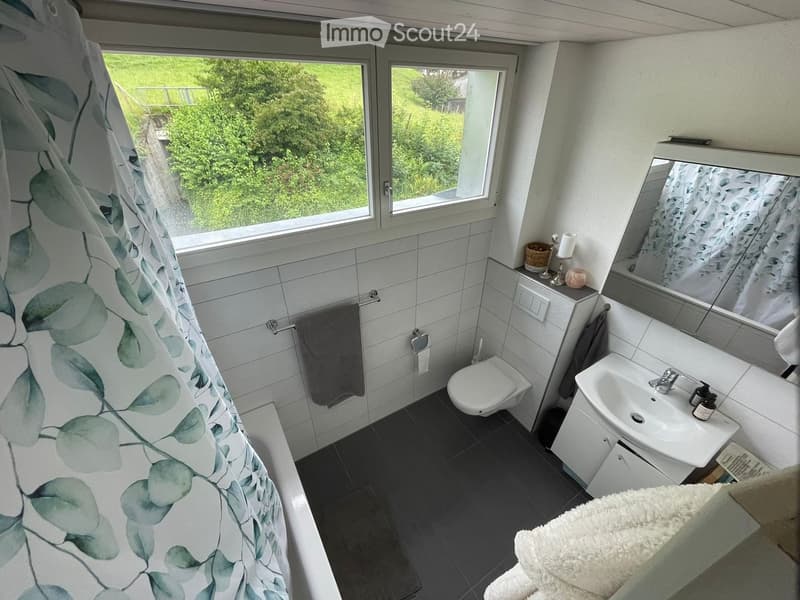 Modern bathroom with white tiles, a toilet, a sink with a mirror, a towel, and a shower curtain with a leafy pattern. The room has large windows offering a view of greenery outside.