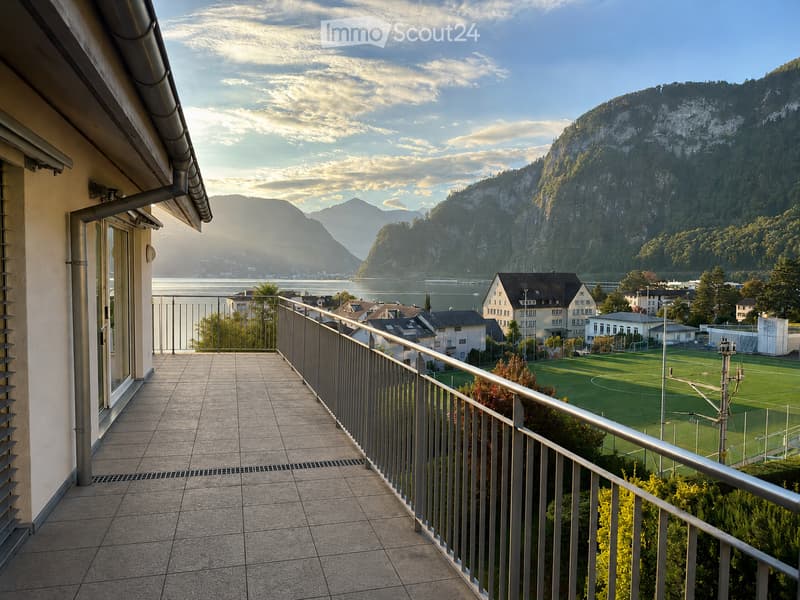 Balcony with tiled flooring, metal railing, overlooking a scenic lake and mountain landscape, with a soccer field in the foreground.