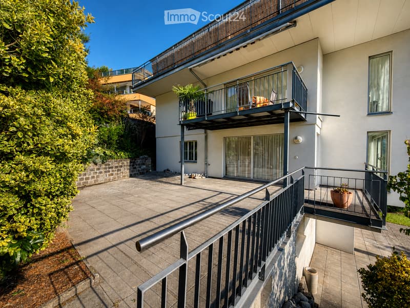 Balcony with metal railing, potted plants, and outdoor furniture, attached to a modern building with large windows and a view of greenery.