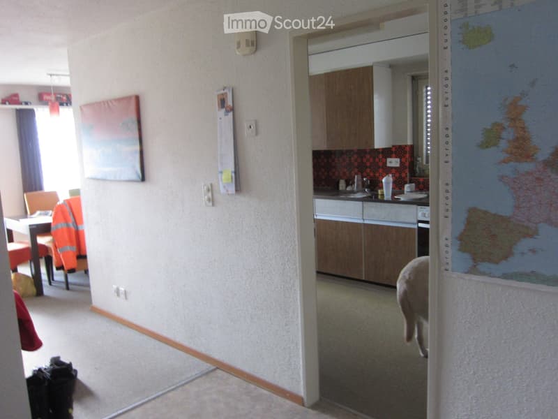 A furnished room with a dining area and a small kitchen. The dining area includes a table and chairs, while the kitchen features cabinets, a sink, and various small appliances.